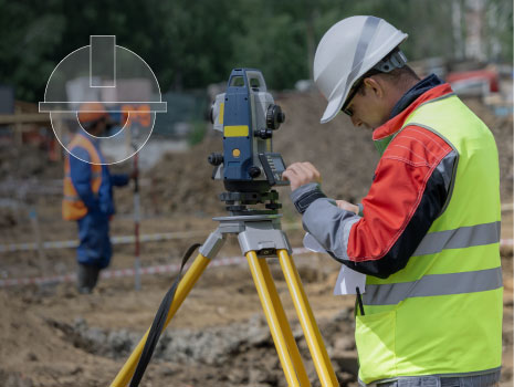 A construction site surveyor wearing a high-visibility jacket and hard hat operates a total station, an advanced surveying instrument mounted on a tripod, to measure distances and angles for precise site mapping. In the background, another worker in safety gear holds a leveling rod, assisting with the surveying process. The site features dirt, construction equipment, and marked areas, emphasizing the importance of accurate measurements in construction planning and execution.